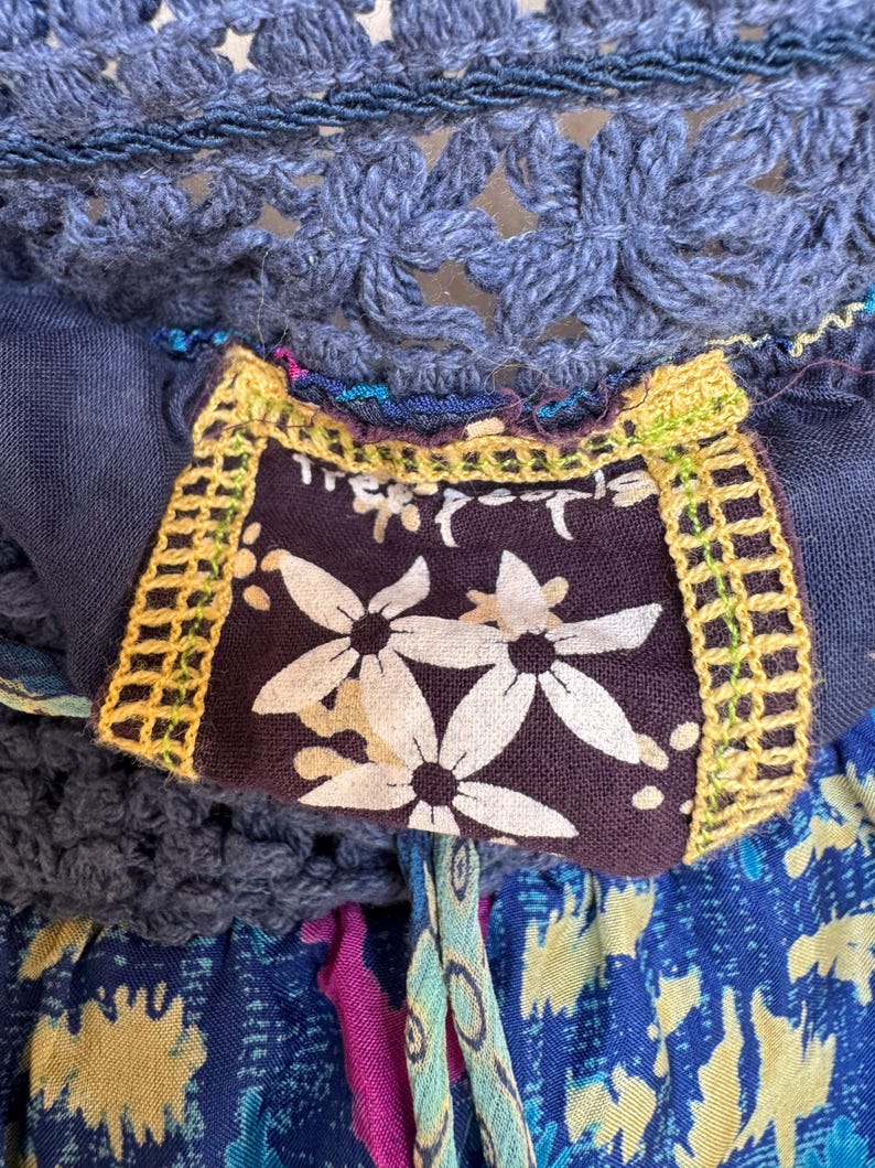 Puede incluir: Primer plano de un top de ganchillo azul con un parche estampado floral. El parche es de color p&uacute;rpura oscuro con flores blancas y ribete amarillo. La parte superior tiene un cord&oacute;n y est&aacute; hecha de varias telas azules.