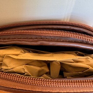 May include: Close-up of a brown leather bag with a zipper closure. The bag is partially open, revealing crumpled brown paper inside. The zipper and leather have a consistent color.