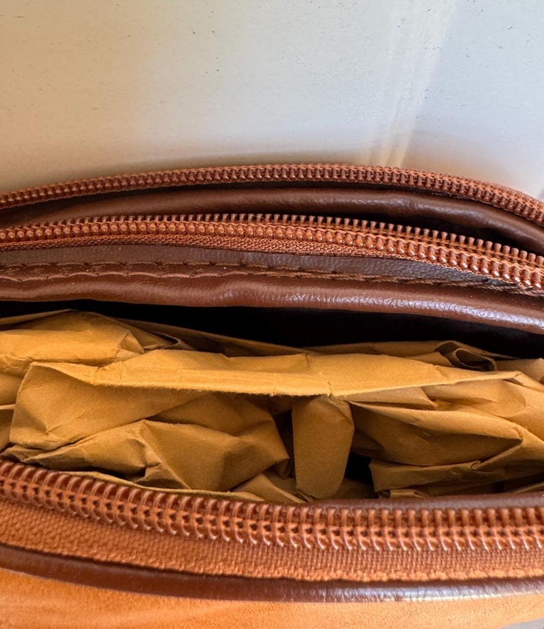 May include: Close-up of a brown leather bag with a zipper closure. The bag is partially open, revealing crumpled brown paper inside. The zipper and leather have a consistent color.