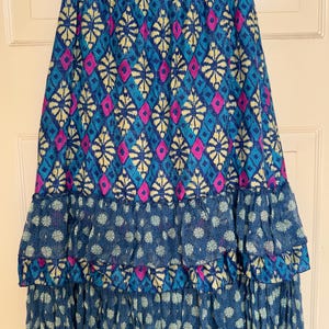Puede incluir: Falda larga con cinturilla de ganchillo azul. La falda presenta un vibrante estampado de diamantes y formas florales en tonos azules, rosas y amarillos. Las capas con volantes en la parte inferior a&ntilde;aden volumen y textura.
