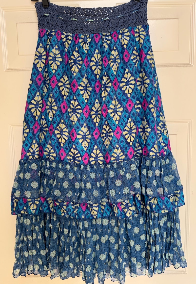 Puede incluir: Falda larga con cinturilla de ganchillo azul. La falda presenta un vibrante estampado de diamantes y formas florales en tonos azules, rosas y amarillos. Las capas con volantes en la parte inferior a&ntilde;aden volumen y textura.
