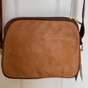 May include: A tan leather crossbody bag with a brown trim and a gold zipper. The bag has a long brown strap and a tag attached. The bag is rectangular with rounded corners.