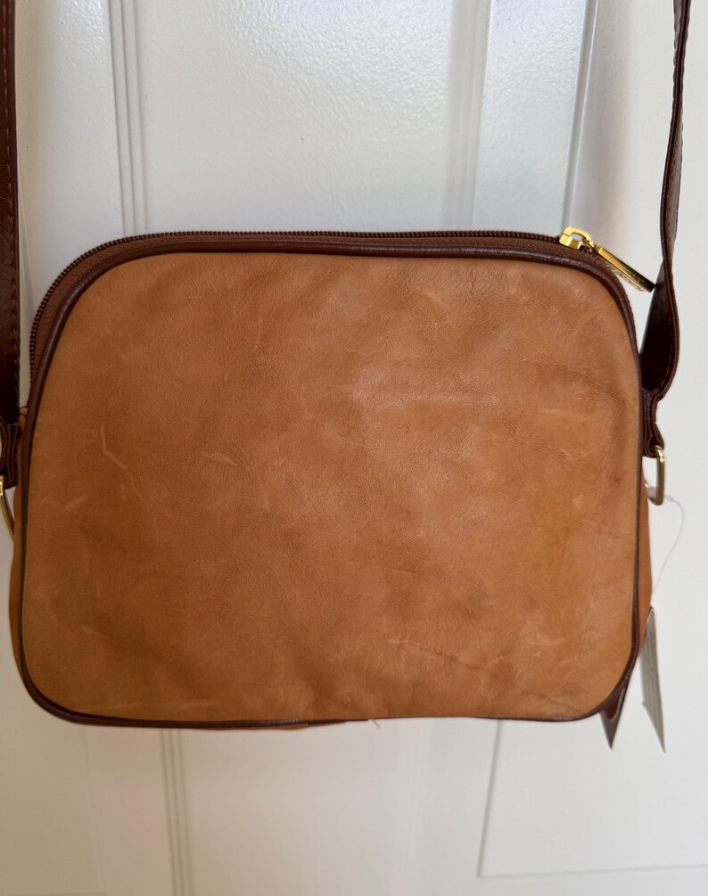May include: A tan leather crossbody bag with a brown trim and a gold zipper. The bag has a long brown strap and a tag attached. The bag is rectangular with rounded corners.