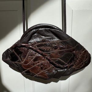 May include: A brown leather handbag with a unique patchwork design. The bag is made of different pieces of leather, some of which have a textured or embossed pattern. The bag has a single strap and a curved, rounded shape.