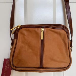 May include: A tan leather crossbody bag with a brown strap and gold-tone hardware. The bag features a vertical zipper and a tag that reads "Valentino di Paolo". The bag is rectangular with rounded corners.