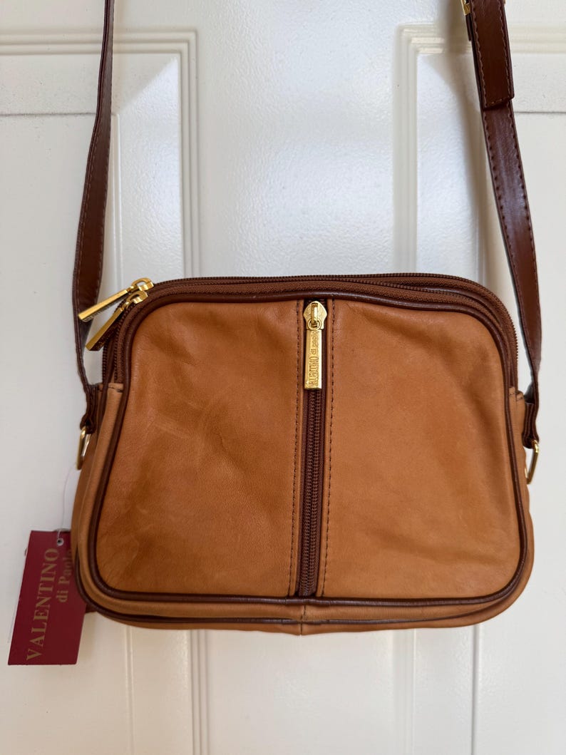 May include: A tan leather crossbody bag with a brown strap and gold-tone hardware. The bag features a vertical zipper and a tag that reads "Valentino di Paolo". The bag is rectangular with rounded corners.