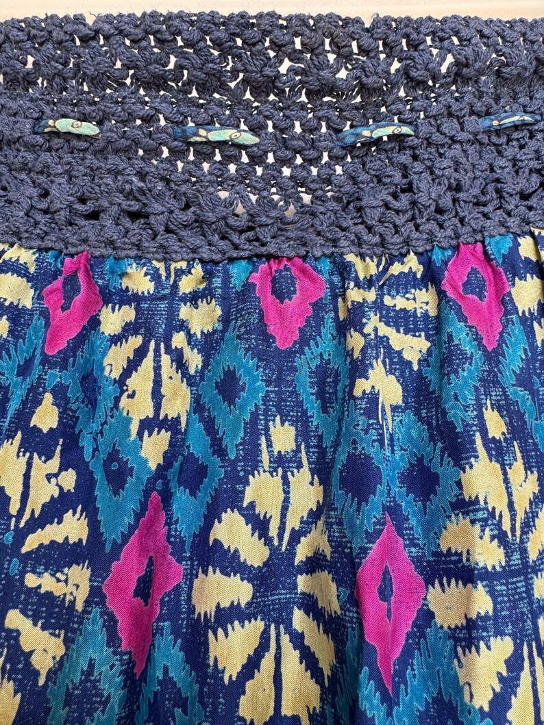 Puede incluir: Primer plano de una falda con una cintura de ganchillo azul marino y una tela estampada. La tela presenta un dise&ntilde;o vibrante con formas de diamantes azules, amarillos y rosas. El dise&ntilde;o de la falda es bohemio y colorido.