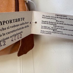 May include: A tan leather bag with a tag attached. The tag has text in multiple languages, including Italian and English, stating that the marks on the bag are characteristic of the handmade process.