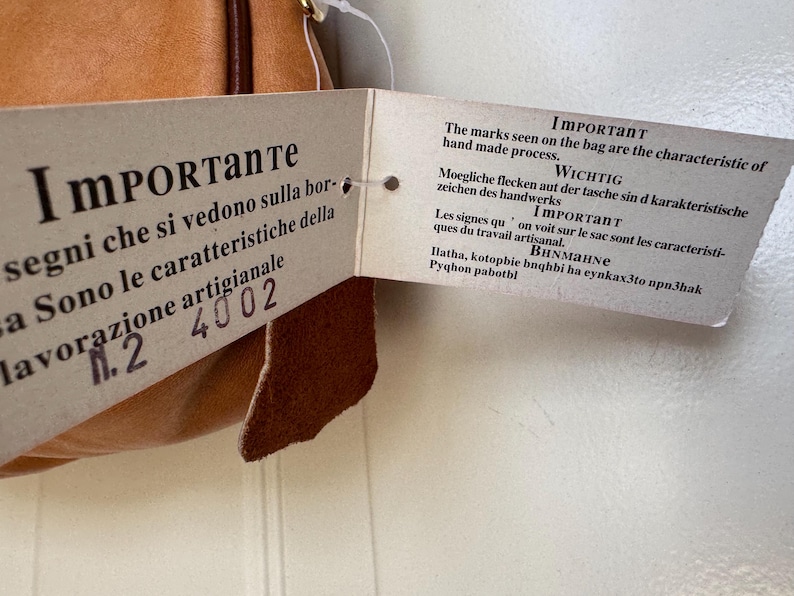 May include: A tan leather bag with a tag attached. The tag has text in multiple languages, including Italian and English, stating that the marks on the bag are characteristic of the handmade process.