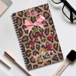 Könnte beinhalten: Spiralgebundenes Notizbuch mit Leopardenmuster. Der Einband zeigt eine rosa Schleife, Kirschen und den Text "Kiss Me I'm a Cherry". Weitere Grafiken sind Lippen, eine Krone und die Worte "Elite Trend".
