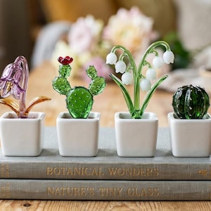 May include: Four glass plant sculptures in white square pots sit atop two books. The plants include a purple flower, a green cactus with a red flower, white lily of the valley, and a green cactus. The books read "Botanical Wonders" and "Nature's Tiny Glass."
