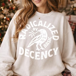 Op de afbeelding: Crèmekleurige sweatshirt met de tekst "RADICALIZED BY BASIC DECENCY" in een cirkelvormig ontwerp. Een witte duif met een takje staat in het midden van het ontwerp. De sweatshirt is neutraal van kleur en heeft een ronde hals.