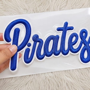 May include: A blue embroidered patch featuring the word Pirates in a cursive script font. The lettering has a thick white border outline and is presented on a clear plastic backing sheet, showcasing the detailed thread texture of the design.