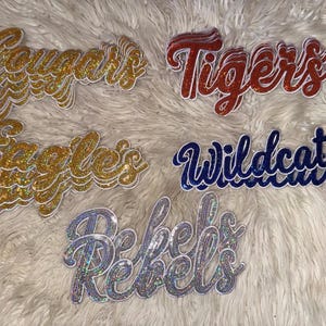 May include: Five sets of layered script word patches featuring glittery finishes. The patches display the words Cougars and Eagles in gold, Tigers in red, Wildcats in blue, and Rebels in a silver holographic finish, each with white borders.