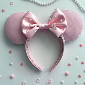 May include: A pink headband with mouse ears and a bow. The ears are a textured pink fabric with white trim. The satin bow is decorated with small white pearls. The headband is surrounded by decorative pearls and heart-shaped embellishments.