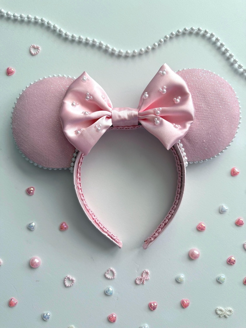 May include: A pink headband with mouse ears and a bow. The ears are a textured pink fabric with white trim. The satin bow is decorated with small white pearls. The headband is surrounded by decorative pearls and heart-shaped embellishments.