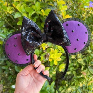 May include: A headband featuring two round purple ears with black polka dots and a central black sequined bow. The ears are edged with black mesh fabric, and the headband itself is covered in a matching black material for a cohesive look.
