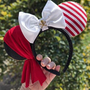 Pode incluir: Uma tiara preta com orelhas com tema de pirata. Uma orelha é listrada em vermelho e branco, a outra é vermelha com uma borda preta. Um grande laço branco com um pingente de caveira e ossos cruzados dourados é centralizado na tiara.
