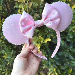 May include: A pink mouse ear headband featuring a satin bow decorated with small pearls. The ears have a glittery texture and a scalloped edge. The headband is held in a hand against green foliage.