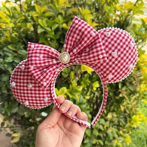 May include: A headband featuring two circular ears and a central bow, all covered in a red and white gingham fabric. The ears and bow are embellished with small white pearls and heart-shaped beads, with a larger heart-shaped charm at the center.
