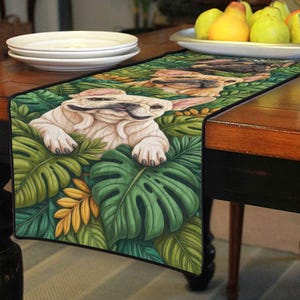 May include: A table runner featuring a whimsical illustration of a French Bulldog nestled among lush green tropical leaves. The runner's design includes a black border and a vibrant color palette, adding a touch of nature-inspired charm to any dining setting.