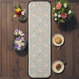 Puede incluir: Una alfombra de pasillo azul claro con un estampado floral y de conejos. La alfombra tiene un borde negro. La imagen también incluye dos platos con comida y tres jarrones con flores.