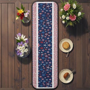 Puede incluir: Un camino de mesa patriótico con fondo azul marino y un patrón de estrellas rojas, blancas y azules. El camino de mesa tiene un borde blanco con detalles de estrellas rojas. Sobre una superficie de madera, con flores y platos.
