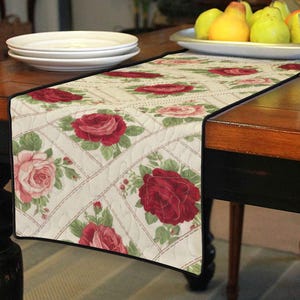 Könnte beinhalten: Ein floraler Tischläufer mit cremefarbenem Hintergrund, gemustert mit roten und rosa Rosen und grünen Blättern. Er hat einen schwarzen Rand und liegt auf einem dunklen Holztisch. Der Läufer ist etwa 152 cm lang.