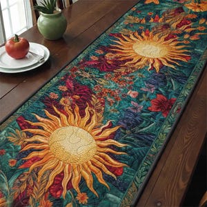 Op de afbeelding: Een gewatteerde tafelloper met een levendig zonontwerp, met gele en oranje stralen tegen een turquoise achtergrond met bloemen- en bladerpatronen. De loper is afgewerkt met een donkergroene rand, geschikt voor elke tafelsetting.