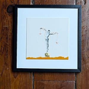 May include: Framed artwork featuring a minimalist figure suspended by strings with red bows. The figure is depicted in black and white, with a gold-colored base and orange accents. The artwork is encased in a black frame.