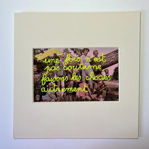 May include: Framed artwork with a vintage photograph and neon green embroidered text. The photograph shows a group of people outdoors. The text reads "une fois n'est pas coutume faisons les choses autrement."