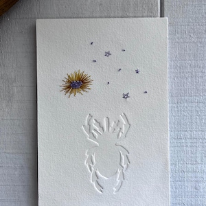 May include: White card with a pressed flower and star accents. The card features a pressed yellow and purple flower, small purple stars, and a debossed insect silhouette. The card is on a light blue wooden surface.