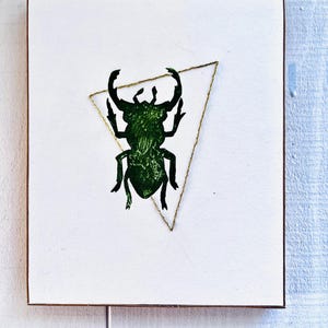 May include: Square artwork featuring a green insect silhouette within a gold triangle on a white background. The artwork has a brown wooden frame. The insect has large, curved antennae. The background is a textured white surface.