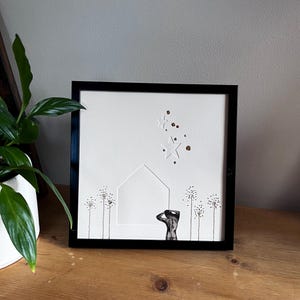 May include: A framed artwork featuring a white background with a cut-out house shape and star accents. The artwork includes a black and gray figure and delicate floral elements. The frame is black, and the piece is displayed on a wooden surface.