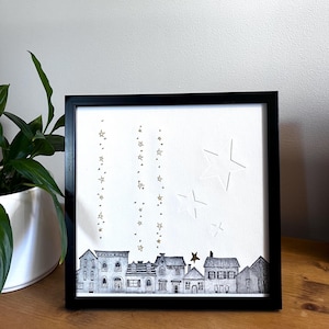 Peut inclure: Impression d'art encadrée en noir représentant un paysage urbain avec des maisons détaillées. Des étoiles dorées et blanches sont dispersées sur le fond blanc. L'œuvre est exposée sur une surface en bois.