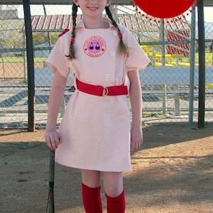 May include: A young person dressed as a baseball player wearing a pink uniform with a red belt, a red baseball cap with a white letter R, red knee-high socks, and black sneakers. The uniform has a patch with a scale design and the text "City of Rockford Peaches". The person is holding a black baseball bat.