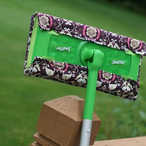 Puede incluir: Un recogedor Swiffer verde con una funda de tela floral. El recogedor está apoyado en un poste de madera.