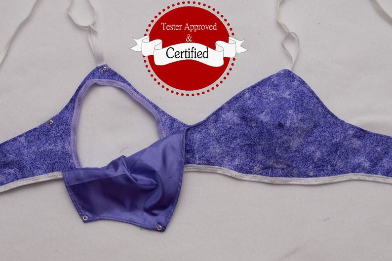 May include: A purple and white fabric bra with a white trim and a purple satin lining. The bra has a white strap on each side. The bra is on a white surface. The text "Tester Approved & Certified" is in a red circle with a white ribbon.