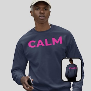 May include: Navy blue sweatshirt with the word "CALM" in bright pink block letters across the chest. A smaller image shows the back of the sweatshirt with the word "CHAOS". The sweatshirt is a casual style.