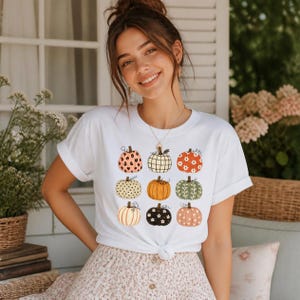 Könnte beinhalten: Ein weißes Kurzarm-T-Shirt mit einem Grafikdruck von neun Kürbissen in verschiedenen Mustern und Farben, darunter Leopardenmuster, Karos, Blumen, Punkte und einfarbiges Orange, angeordnet in einem drei-mal-drei Raster auf der Vorderseite.