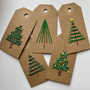 May include: Five rectangular brown kraft paper tags featuring hand-stitched embroidery designs of Christmas trees. Each tag has a circular hole at the top. The trees are created with green and brown thread, with some featuring small colorful accents.