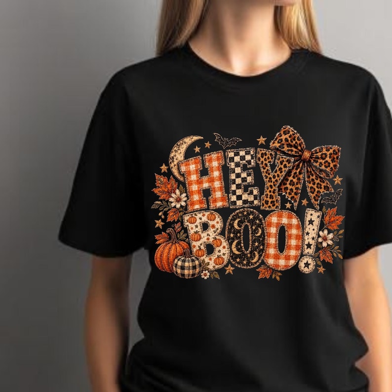 K&ouml;nnte beinhalten: Ein schwarzes Kurzarm-T-Shirt mit einem Grafikdruck, der in gro&szlig;en Buchstaben HEY BOO zeigt. Das Design umfasst orangefarbene Karomuster, schwarz-wei&szlig; karierte Muster, Leopardenmuster sowie Akzente mit K&uuml;rbissen, Sternen und einer gro&szlig;en Leoparden-Schleife.