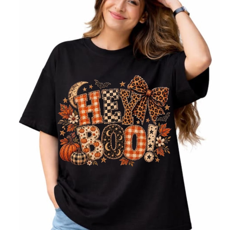 K&ouml;nnte beinhalten: Ein schwarzes Kurzarm-T-Shirt mit dem Schriftzug HEY BOO in gro&szlig;en Blockbuchstaben mit orangefarbenen Karomustern, schwarz-wei&szlig;em Schachbrettmuster und Leopardenmuster. Das Design enth&auml;lt K&uuml;rbisse, Herbstbl&auml;tter, Sterne, Flederm&auml;use und eine gro&szlig;e Schleife.
