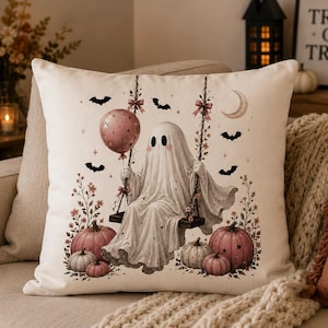 Könnte beinhalten: Ein quadratisches Dekokissen mit einem weißen Geist auf einer Schaukel, der einen rosa Luftballon hält. Das Motiv zeigt schwarze Fledermäuse, eine Mondsichel, rosa Blumenakzente sowie rosa und weiße Kürbisse auf cremefarbenem Grund.