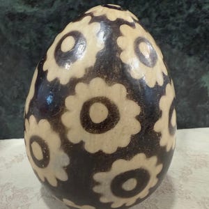 May include: A decorative, egg-shaped object with a dark brown background and cream-colored floral patterns. The egg is likely ceramic or pottery, and the floral design is evenly spaced across the surface.