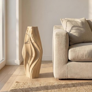 Pode incluir: Um vaso de chão alto, de cor bege, com uma textura ondulada de nervuras verticais. O vaso apresenta uma silhueta angular e afunilada com um acabamento mate suave, posicionado verticalmente sobre um chão de madeira clara junto a um sofá.
