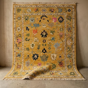 May include: Mustard yellow area rug with a floral pattern in shades of pink, blue, green, and orange. The rug has a cream-colored border and fringe along the edges. The rug is displayed on a neutral-colored floor.
