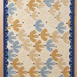 Puede incluir: Alfombra rectangular con un patrón repetido de pájaros estilizados en tonos crema, beige y azul. La alfombra tiene un borde azul oscuro con un diseño en forma de ojo. Los pájaros están dispuestos en filas sobre un fondo crema.
