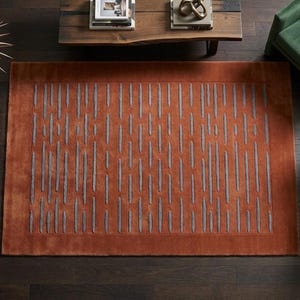 May include: A rectangular, rust-coloured rug featuring a pattern of vertical grey lines. The rug is placed on a dark wood floor, with green chairs and a wooden table in the background. The rug measures approximately 152 cm by 228 cm.