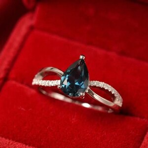 May include: A silver ring featuring a dark blue, pear-cut gemstone. The band splits, each side adorned with small, clear stones. The ring is set against a red velvet background.